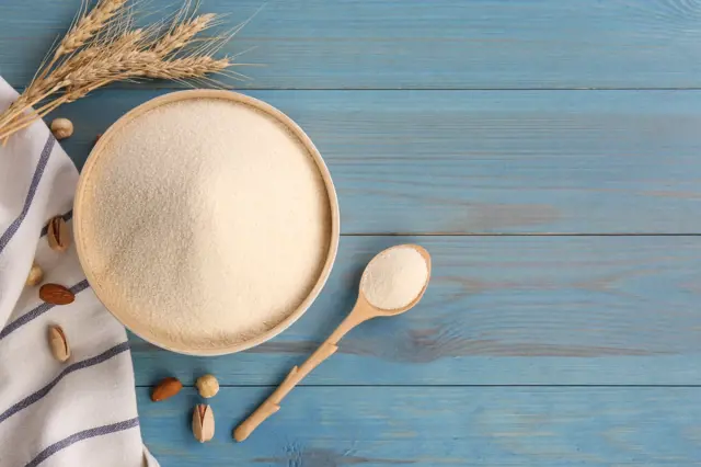 Semolina v dřevěné mísce s pšenicí a ořechy na modrém pozadí.