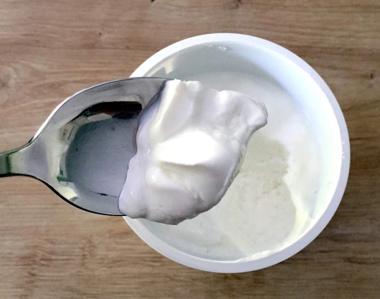Lžíce s řeckým jogurtem nad otevřenou nádobkou na dřevěném povrchu.