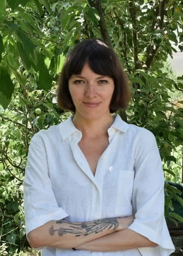 Žena s tetováními na pažích stojí venku před zelenou vegetací.
