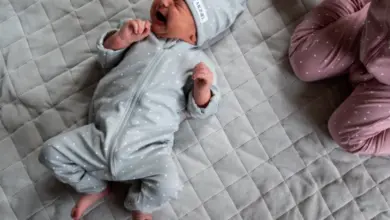 Pláčající miminko v šedém pyžamu leží na posteli.