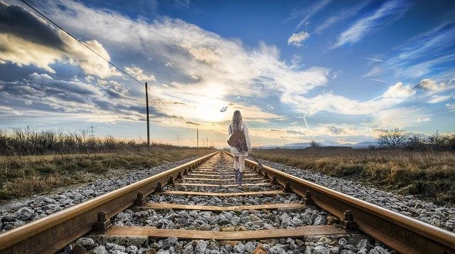 Žena kráčí po železničních kolejích k slunci pod mraky.