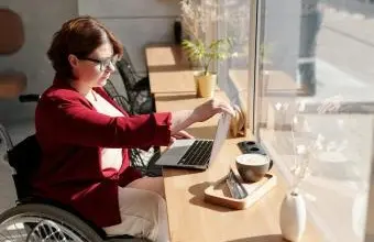Žena v invalidním vozíku pracuje na notebooku u okna.