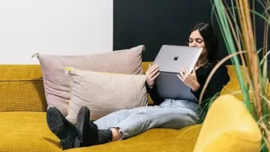 Žena sedí na žluté pohovce s notebookem v klidném prostředí.