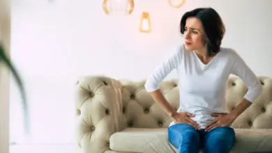 Žena sedí na gauči s rukama na břiše v bolesti.