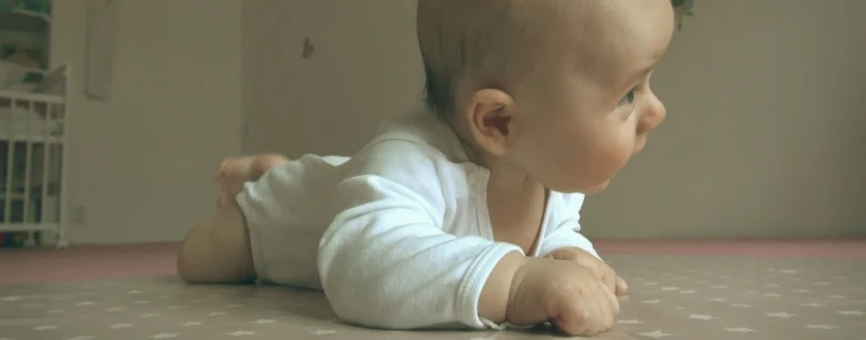 Miminko v bílé košilce leží na bříšku na hrací koberci.