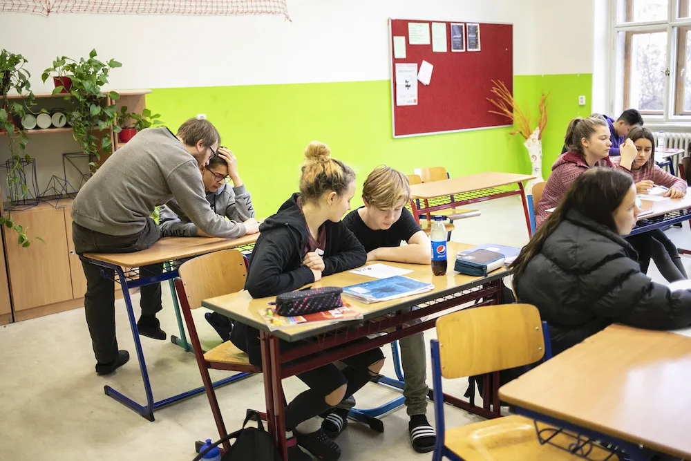 Školní třída s učiteli a žáky sedícími za stoly.