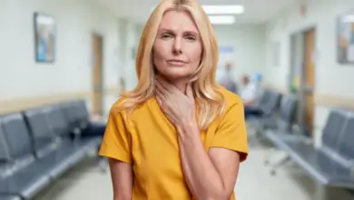 Žena v nemocniční chodbě drží ruku na krku po operaci štítné žlázy.