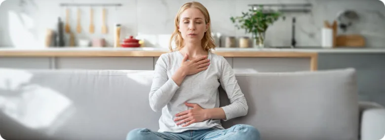 Žena sedí na gauči s rukama na hrudi při meditaci pro úzkost.