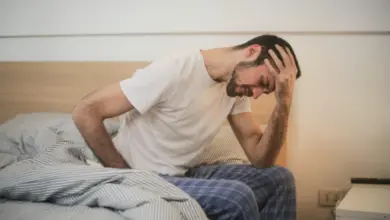 Muž sedí na posteli a drží si hlavu kvůli bolesti zad.