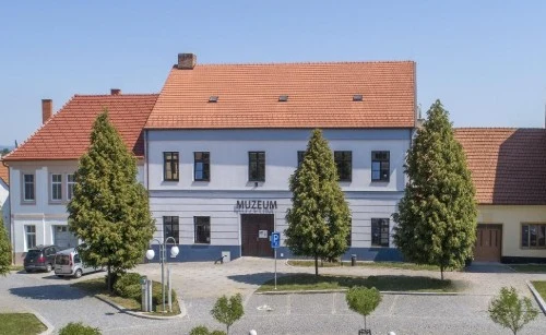 Budova muzea v Mikulčicích s červenou střechou a okolní zeleň.