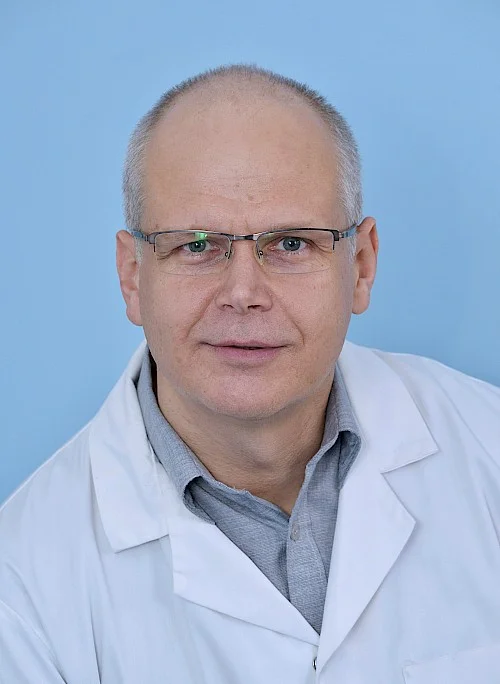 Muž v bílém lékařském plášti s brýlemi na modrém pozadí.