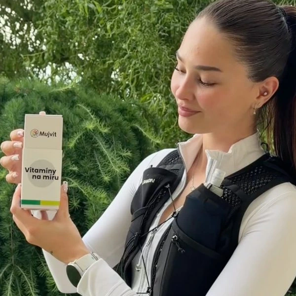 Žena drží krabičku vitamínů na pozadí zelené vegetace.